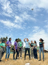 Students stand around in front of the farm where the field school takes place, watching a flying drone.